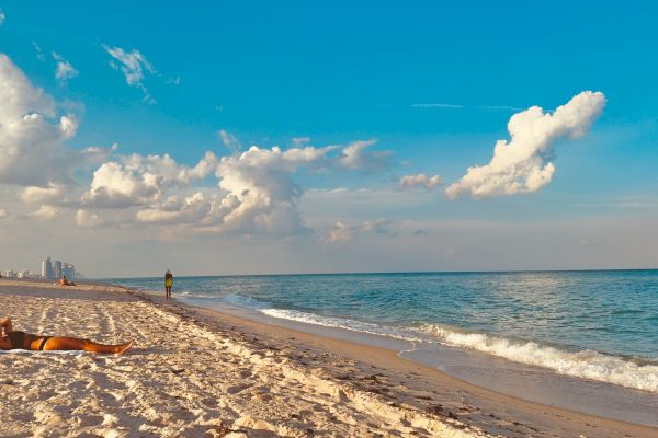 A person sunbathes on a sandy beach with ocean waves, blue sky, and clouds. City buildings are visible in the distance.