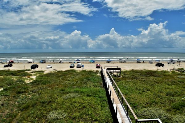 A beach scene with cars, tents, and people along the shoreline, blue sky, clouds, and a wooden path over green dunes leading to the sand.