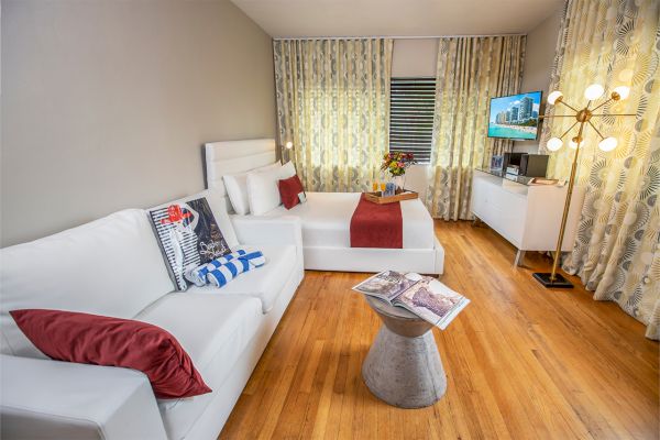 A modern living room with a white sofa, colorful cushions, a coffee table with a book, a bed with a red throw, and a TV.