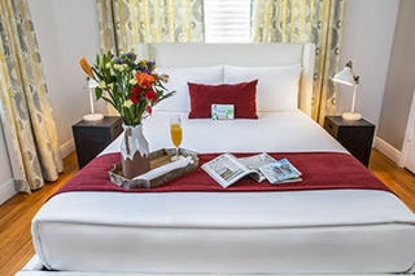 A neatly made bed with a red runner, tray with flowers, magazines, a drink, and two bedside lamps in a well-lit room.