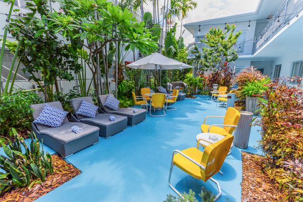 Outdoor patio with blue flooring, gray loungers, yellow chairs, greenery, and a white umbrella. Vibrant and relaxing setting.