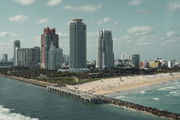 A beach scene with high-rise buildings in the background, a pier stretching into the ocean, and people on the sandy shore under a clear sky.