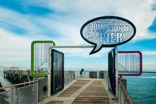 A wooden walkway leads to South Pointe Park Pier, with colorful frames and a speech bubble sign, set against the ocean and blue sky.