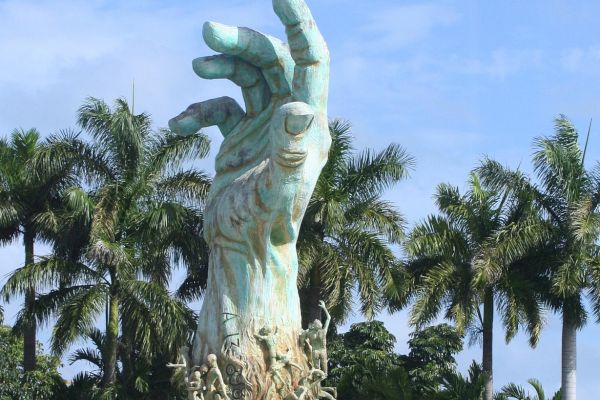 A large hand sculpture with figures climbing its surface, surrounded by palm trees under a blue sky.