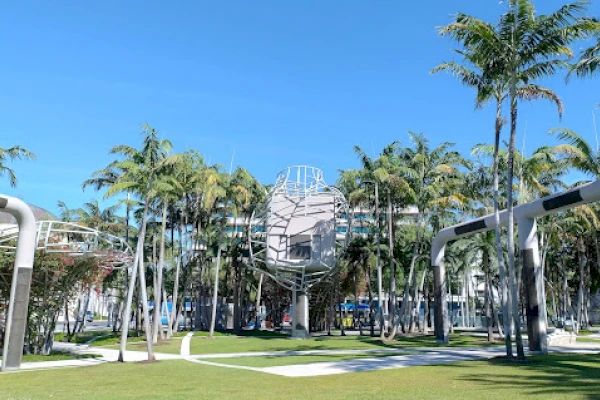 The image shows a park with palm trees, modern sculptures, and a clear blue sky. The scene is tranquil and landscaped.