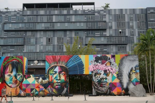 The image shows a colorful mural featuring diverse faces, set against a modern, geometric-patterned building backdrop.