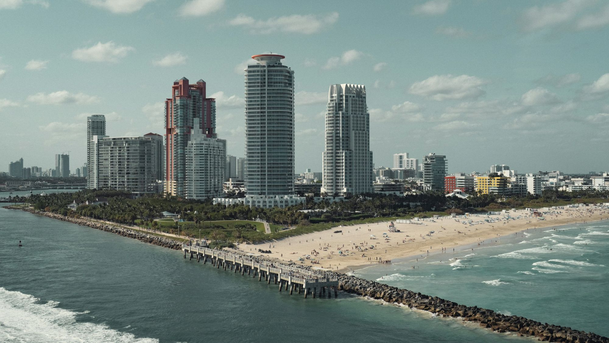 A coastal cityscape with tall buildings, a sandy beach, and a pier extending into the ocean, set under a partly cloudy sky.