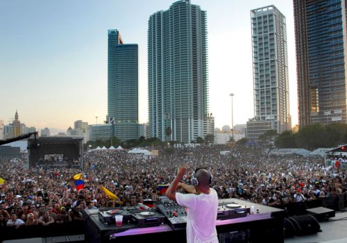 A DJ performs on stage in front of a massive crowd, with tall city buildings in the background at sunset during a lively outdoor concert.